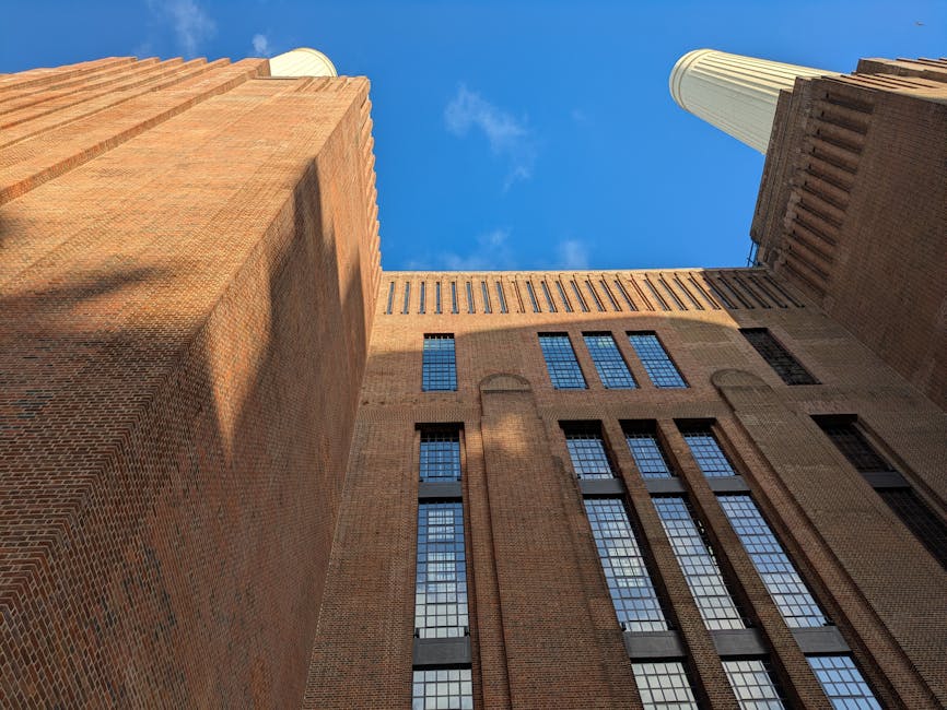 Looking up at tall brick buildings and skyscrapers in Battersea during daytime, with clear blue sky visible above. The structures feature large, rectangular windows with black frames, some of which ar