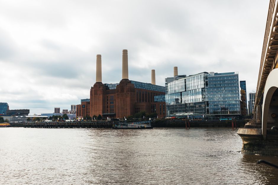 View of Battersea Power Station from the river, with three tall brick chimneys and modern glass-fronted buildings nearby. In the foreground, the river reflects the cloudy sky above, and part of a bridge or loading area is visible on the right side. Inside a property close to the river, cardboard boxes, wrapped furniture, and packing materials are seen being prepared for a home relocation. A trolley or moving straps might be visible as part of the furniture transport process. The scene captures the loading or packing stage involved in a professional removals service by Battersea Movers, highlighting the urban environment and the logistics of packing and moving within a city setting.