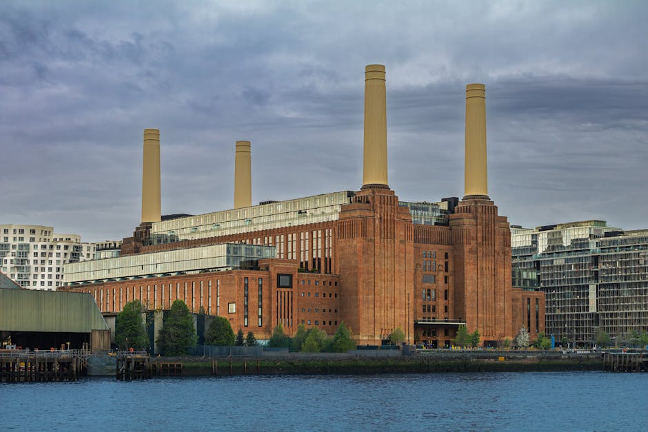 A large, historic building of red brick and concrete situated along a riverbank, with four tall, yellowish industrial chimneys extending from its roof. The building features multiple levels, with sections of glass windows and modern extensions, and is surrounded by green trees and a paved outdoor area. In the foreground, there is a wooden pier or loading dock extending into the water, with parts of a moving van or transport vehicle visible nearby, suggesting active loading or unloading processes. The sky above is overcast with grey clouds, indicating a typical day of home relocation or furniture transport involving careful handling of packed boxes and furniture. This scene reflects the logistics involved in house removals near Battersea Power Station, as exhibited by Battersea Movers, and illustrates the logistics of packing and moving goods efficiently in urban environments.