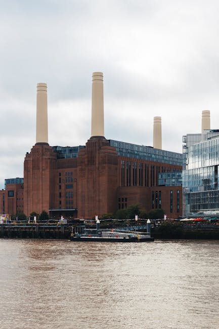 View of Battersea Power Station from the river, with three tall brick chimneys and modern glass-fronted buildings nearby. In the foreground, the river reflects the cloudy sky above, and part of a bridge or loading area is visible on the right side. Inside a property close to the river, cardboard boxes, wrapped furniture, and packing materials are seen being prepared for a home relocation. A trolley or moving straps might be visible as part of the furniture transport process. The scene captures the loading or packing stage involved in a professional removals service by Battersea Movers, highlighting the urban environment and the logistics of packing and moving within a city setting.