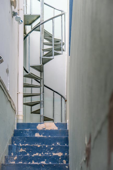Interior view of a narrow staircase with worn blue-painted steps, showing peeling paint and signs of usage. The staircase has a metal spiral structure with a curved dark railing, leading upward from a small landing area. To the left, there is a white wall with visible pipes and a door frame, while to the right, a plain wall partially visible. The lighting appears natural, illuminating the space evenly. At the base of the stairs, there are several cardboard boxes and a wrapped piece of furniture, indicating ongoing packing or home relocation activities. The setting suggests a typical residential building where careful packing and furniture transport are necessary to navigate the tight staircase during a move, with Battersea Movers potentially providing assistance with such house removals involving narrow staircases.