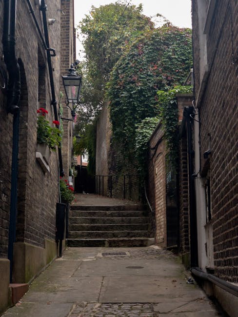 A narrow outdoor alleyway between two brick buildings, featuring a series of stone steps leading upward towards a black metal gate. The alley is paved with concrete and bordered by brick walls on both sides, with black metal railings on the left side supporting potted plants and flowers, including red blossoms. Vegetation, including a large bush with dense green and reddish leaves, partially covers the upper section of the wall on the right. A traditional black lamp is mounted on the building on the left, and the scene is lit with natural daylight, highlighting the textures of the bricks, stone steps, and foliage. This setting is typical for urban residential areas, where home relocation and furniture transport may involve navigating narrow staircases and tight outdoor spaces, features that Battersea Movers is experienced in handling during home removals.