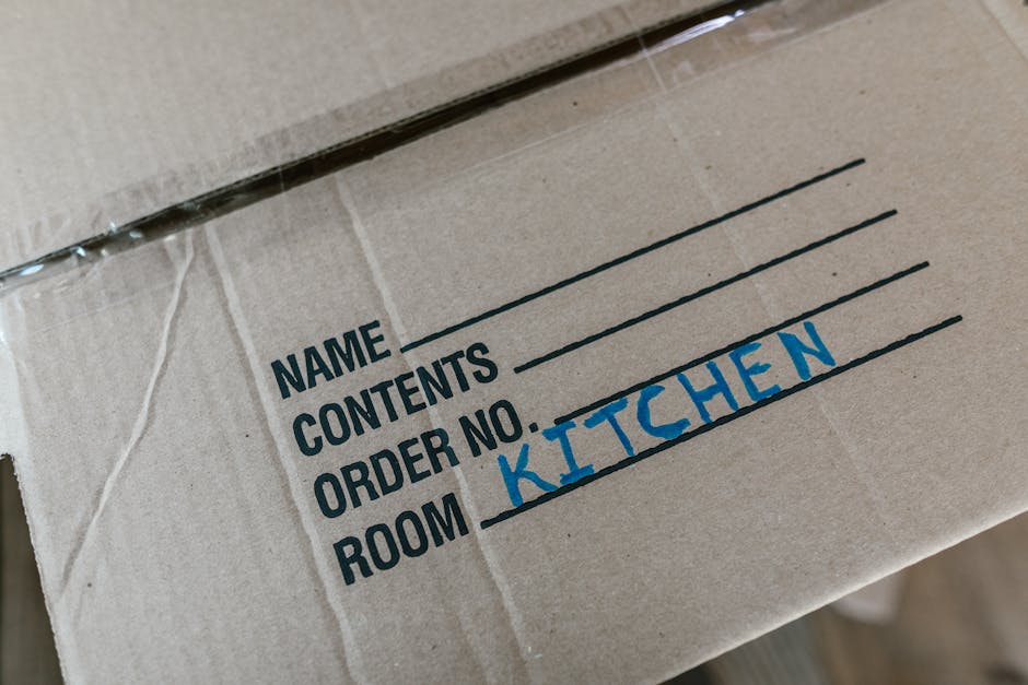 Close-up of a cardboard moving box labeled 'Kitchen' with blue handwritten text, placed amidst other packed boxes on a dolly or trolley. The box displays printed sections for name, contents, order num