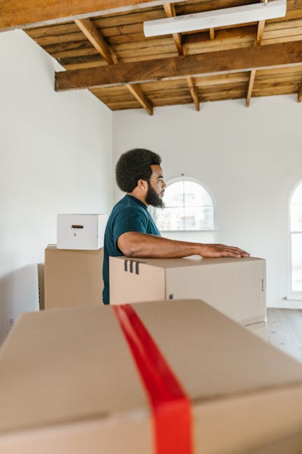 Close-up of a cardboard moving box labeled 'Kitchen' with blue handwritten text, placed amidst other packed boxes on a dolly or trolley. The box displays printed sections for name, contents, order num